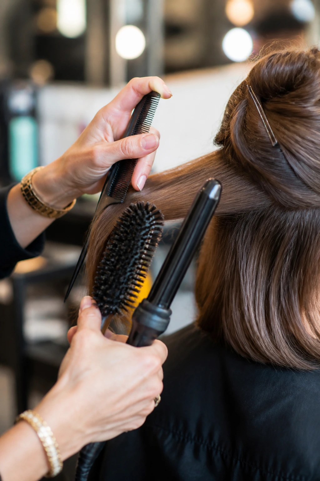 Hands with hair tools (tail comb, brush, curling wand) styling hair in a salon