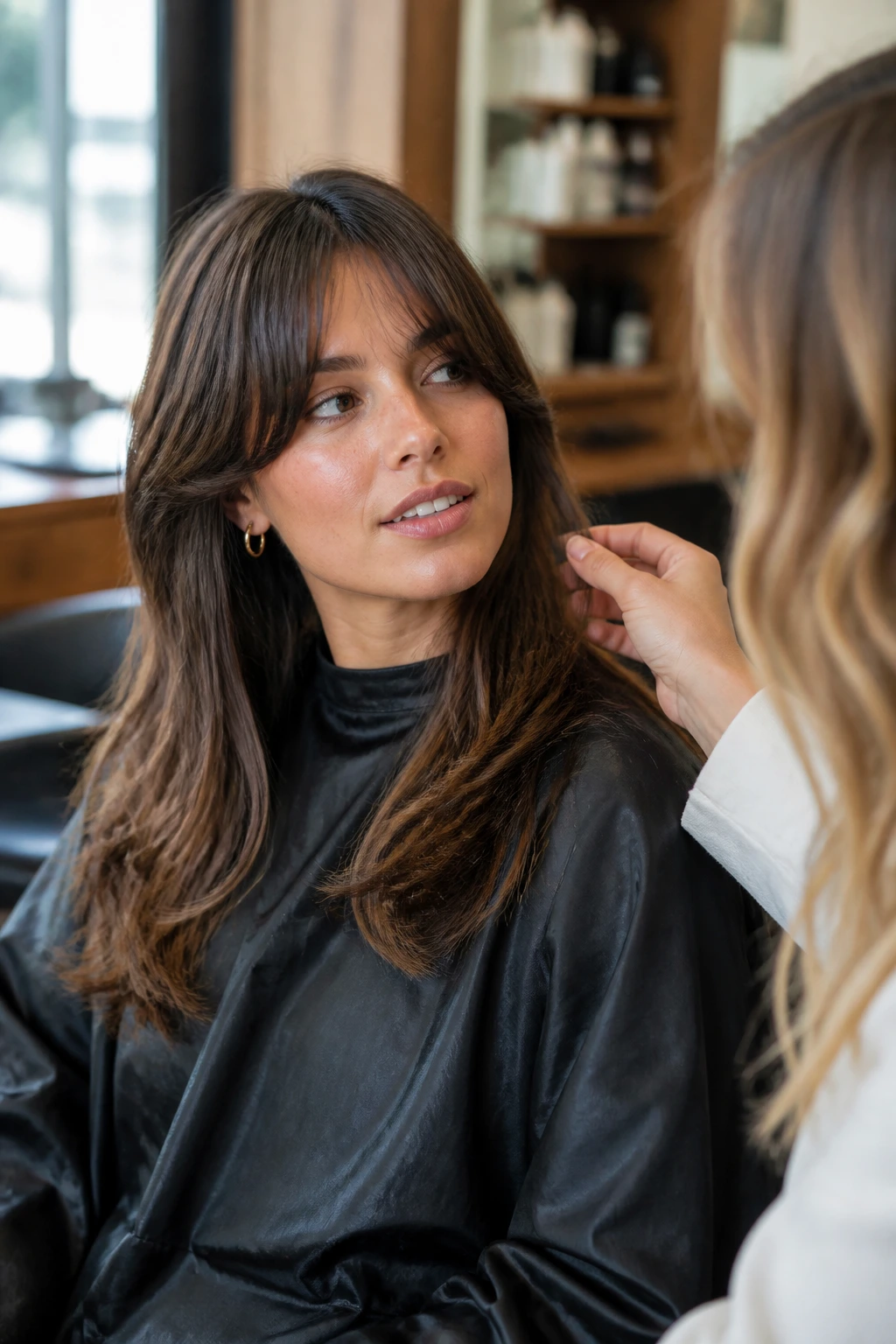 Real woman in a salon chair discussing fringe length with a stylist, warm window light