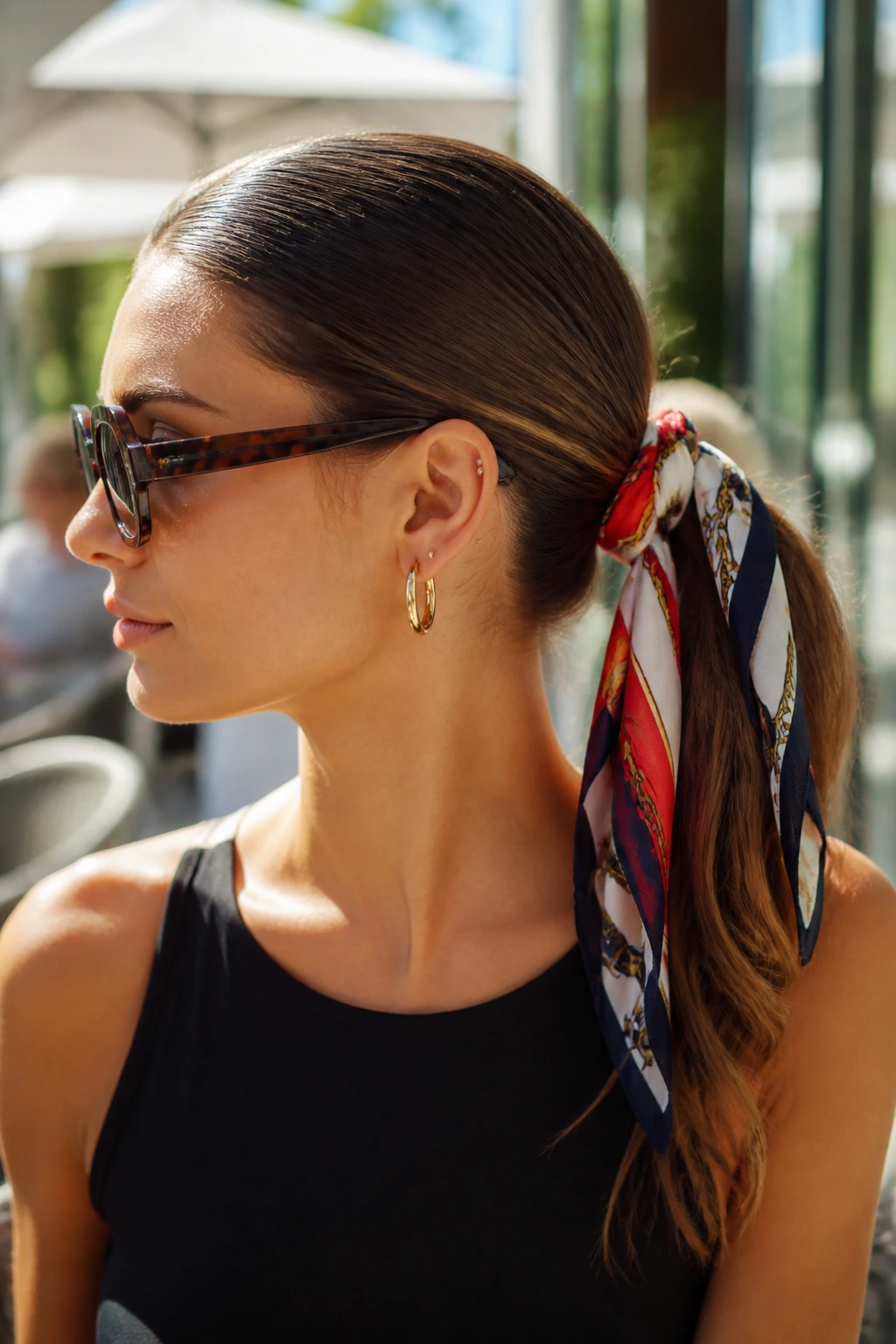 Close-up of a woman with a scarf-wrapped ponytail in an outdoor cafe setting
